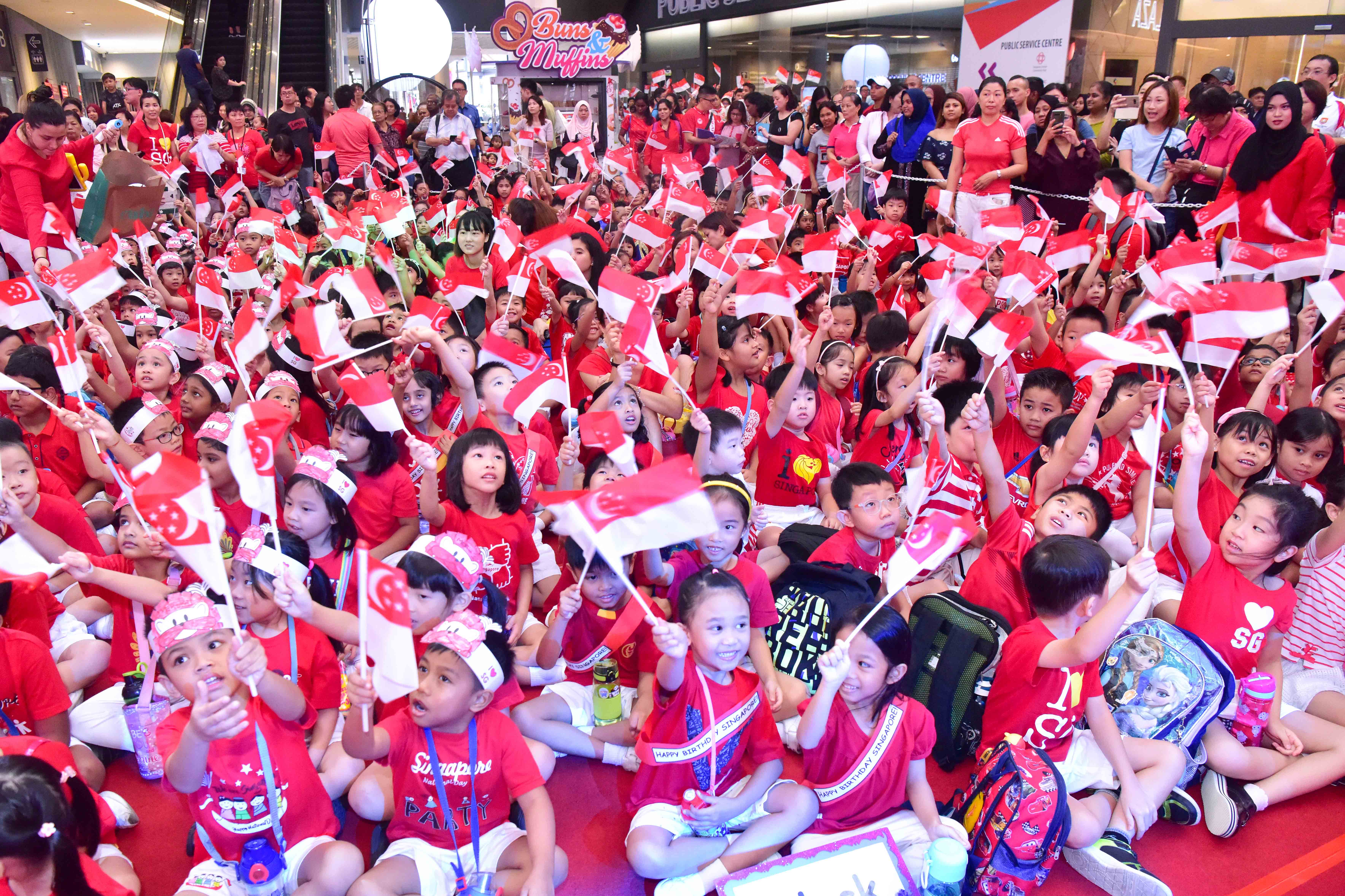 A large group of people, mostly children, wearing red shirts and holding Singapore flags in a crowded indoor event.